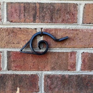 May include: A black metal decorative wall hanging in the shape of a stylized eye, mounted on a red brick wall. The eye has a spiral and a straight line extending from it. The brick wall has white mortar.