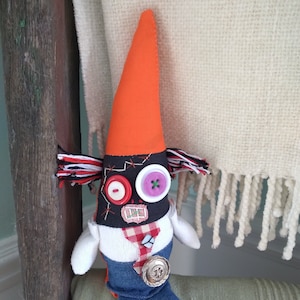 May include: A handmade doll with a black and white striped hat, orange cone hat, and blue jeans. The doll has a black face with red and purple buttons for eyes and a red button for a nose.