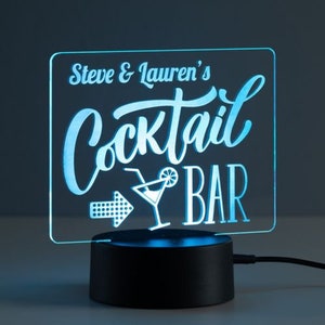 May include: A personalized LED sign for a cocktail bar. The sign is rectangular with the words "Steve & Lauren's Cocktail Bar" in a light blue, illuminated font. A cocktail glass and arrow graphic are also included. The base is black.