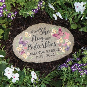 May include: A gray stone garden memorial with the text "Now She Flies with Butterflies" and the name "Amanda Parker" with the dates "1935-2022". The stone is decorated with colorful butterflies and flowers.