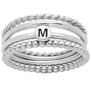 May include: A stack of silver rings with different textures. One ring has a twisted rope design, another has small beads, and a third ring features the letter "M" in black on a white background. The rings are stacked together.
