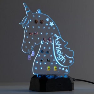 May include: A light blue unicorn-shaped earring holder with a nameplate that reads "Ashleigh". The holder has a black base with a light blue LED light.