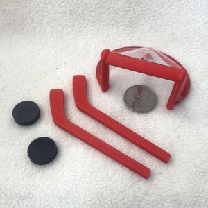 Puede incluir: Una portería de hockey roja en miniatura con red blanca, dos palos de hockey rojos y dos discos negros. Una moneda plateada está colocada junto a la portería para mostrar la escala y el pequeño tamaño de estos accesorios de plástico.