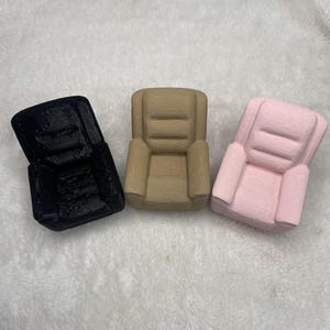 May include: Three miniature armchairs in black, tan, and pink. Each chair has a rectangular back and armrests, with a simple, blocky design. The chairs are arranged on a white, textured surface.