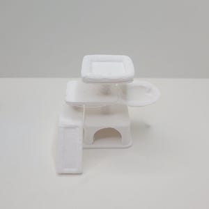 Könnte beinhalten: Ein weißer, 3D-gedruckter Kratzbaum mit mehreren Ebenen. Die Struktur umfasst eine quadratische obere Plattform, eine rechteckige mittlere Ebene, eine gebogene Plattform und eine kleine Nische. Das Design ist minimalistisch und modern, geeignet für kleine Haustiere.