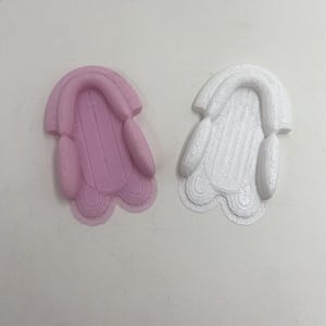 May include: Two 3D-printed objects, one pink and one white, are displayed against a white background. Each object has a rounded top and a rectangular base with rounded edges. The pink object has a smooth surface, while the white one has a textured surface.