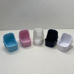 May include: Five miniature baby carriage toys in a row. The carriages are in various colors: light blue, pink, clear, black, and white. Each carriage has a rounded shape with a small interior.