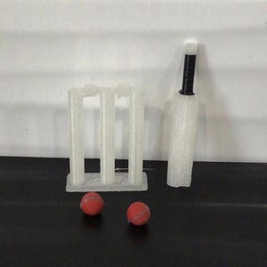 May include: A white 3D-printed cricket wicket set with three stumps and two bails, a matching white cricket bat with a black handle, and two small red cricket balls. The set is arranged on a flat surface, showcasing the textured finish of the items.