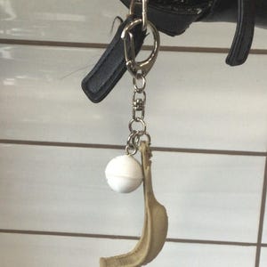 Op de afbeelding: Een sleutelhanger met een witte bal en een bruin banaan-sierlijk. De sleutelhanger is bevestigd aan een zwarte tas met een zilveren rits en een zilveren sluiting.