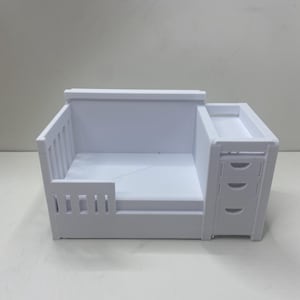 May include: A white, 3D-printed dollhouse crib with a built-in changing table. The crib features a slatted side and a solid headboard. The changing table has three small drawers. The entire piece is made of white plastic.