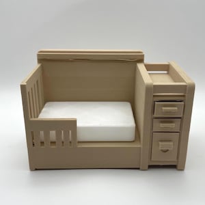 May include: A miniature beige cot with a white mattress and an attached side unit featuring three small drawers. The structure is made of a solid, light-coloured material with a slatted side rail and a flat top surface on the storage section.