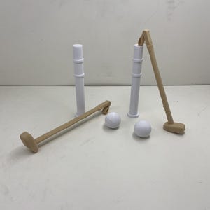 May include: A miniature croquet set with two white posts, two beige mallets, and two white balls. The mallets and posts are arranged on a white surface, suggesting a game setup. The beige mallets have a rectangular head.