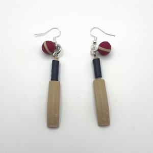 May include: A pair of dangle earrings featuring miniature cricket bats with light tan blades and black handles, paired with small red cricket balls that have a white seam detail. The charms hang from silver-toned metal earring hooks.
