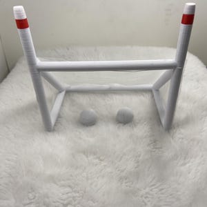 May include: A white, miniature soccer goal with red stripes at the top of each post. Two small, white, spherical objects are positioned in front of the goal. The goal is set on a white, fluffy surface.