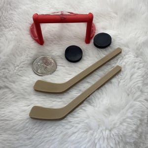 May include: Miniature hockey set featuring a red goal, two black pucks, two tan hockey sticks, and a coin. The set is designed for play or display. The goal has a red net.