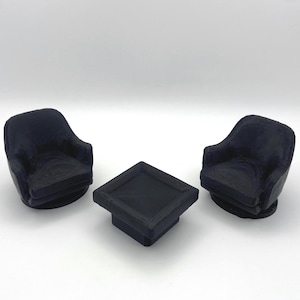 May include: A set of miniature furniture consisting of two black armchairs with curved backrests and a central square table. The items feature a visible layered texture consistent with 3D printing, with a matte finish and a solid black color throughout.
