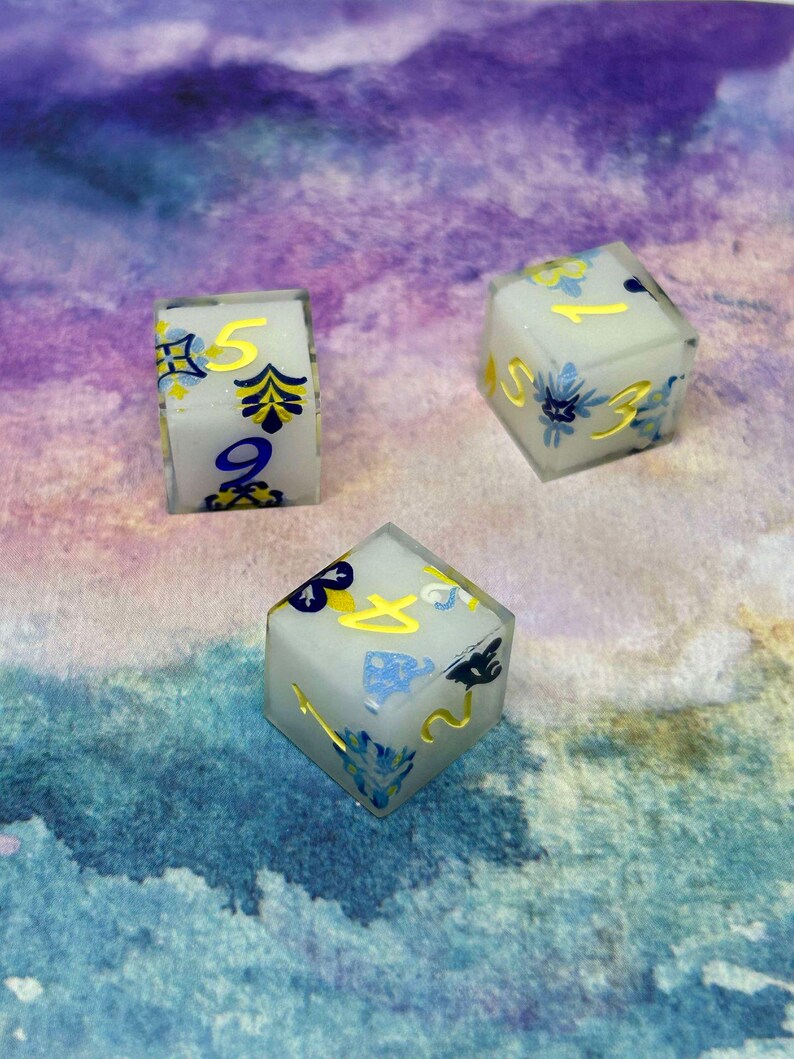 May include: Three white dice with blue and yellow floral designs and gold numbers. The dice are on a purple and blue watercolour background.