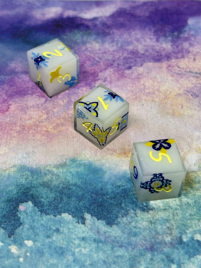May include: Three white dice with blue and yellow floral designs and gold numbers. The dice are on a purple and blue watercolour background.