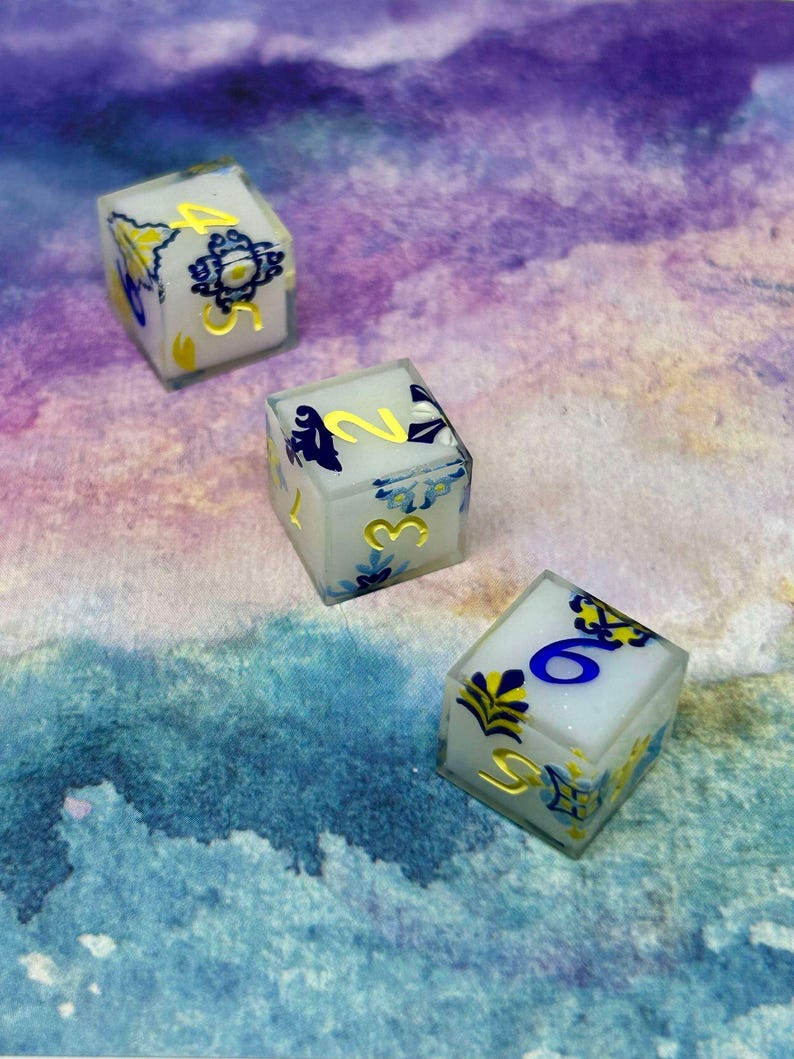 May include: Three white dice with blue and gold designs. The dice are numbered 1, 2, and 6. The dice are on a purple and blue watercolour background.