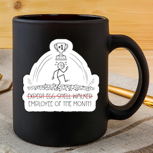May include: Black ceramic mug with a white sticker featuring a stick figure, a trophy, and the text "EXPERT EGG SHELL WALKER EMPLOYEE OF THE MONTH". The mug has a black handle and a glossy finish.