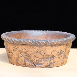 May include: A brown ceramic planter with a textured rim and embossed design. The planter has a wide, shallow bowl shape and is suitable for small plants or succulents. The exterior features a detailed relief pattern.