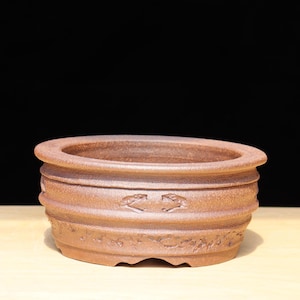 May include: A brown, round bonsai pot with a textured surface. The pot has a wide rim and horizontal ridges around the body. The reddish-brown clay pot sits on a light-coloured surface. The pot is suitable for bonsai trees.