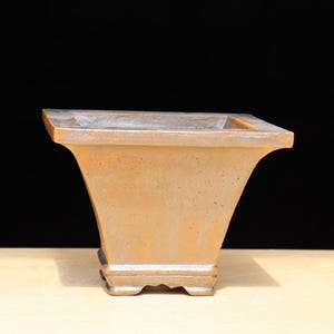 May include: A square, light brown bonsai pot with a flared, trapezoidal shape. The pot has a textured surface and sits on four small feet. The background is black, and the pot is on a light-colored wooden surface.