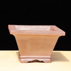 May include: A square, terracotta-colored bonsai pot with a flared shape. The pot has a wide, flat rim and a decorative base. The background is black, and the pot sits on a light-colored surface. This pot is designed for bonsai trees.