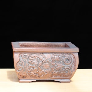 Puede incluir: Una jardinera rectangular de cerámica marrón rojizo con diseños florales y de volutas en relieve. La jardinera tiene una forma ligeramente curvada y una abertura rectangular en la parte superior. El fondo es negro.