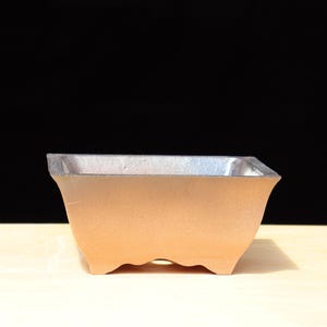 Puede incluir: Una maceta cuadrada de bonsái marrón claro con borde plateado. La maceta tiene un diseño curvo en los lados y una superficie texturizada. La maceta está sobre una superficie marrón claro con fondo negro.