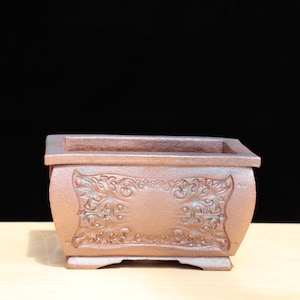 Puede incluir: Una maceta de bonsái rectangular marrón con tallados florales ornamentados en la parte delantera. La maceta tiene una forma ligeramente curvada y un borde elevado. La maceta está hecha de un material marrón rojizo.