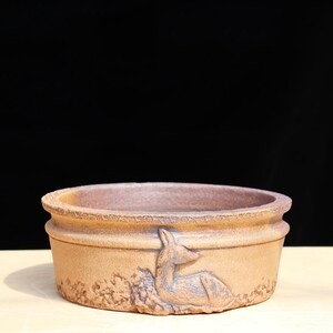 May include: A brown ceramic planter with a deer relief design. The round planter has a textured surface and a decorative band around the middle. The deer is depicted lying down. The planter sits on a light-colored surface against a black background.