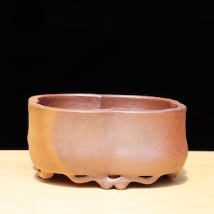 Puede incluir: Una pequeña maceta rectangular de cerámica marrón con esquinas redondeadas y un borde ligeramente ensanchado. La maceta tiene una superficie texturizada y pies decorativos. El fondo es negro.