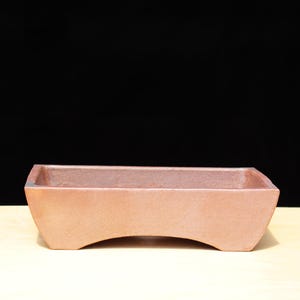 Puede incluir: Una maceta rectangular para bonsái de color terracota, con base curva y lados rectos. La maceta tiene un diseño simple y minimalista y probablemente está hecha de cerámica. El fondo es negro.