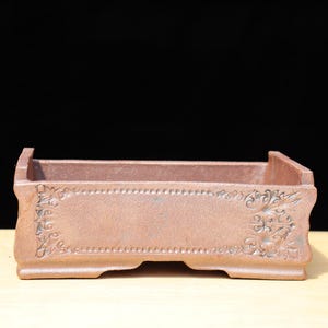 Puede incluir: Una maceta rectangular de color marrón rojizo para bonsái con un diseño floral decorativo en los laterales. La maceta tiene un perfil bajo con un borde ligeramente elevado y está hecha de cerámica. Está diseñada para plantar árboles o arbustos pequeños.
