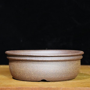 May include: A brown, round bonsai pot with a textured surface. The pot has a wide, shallow design with a raised rim and a small drainage hole. The pot is made of a natural material and sits on a wooden surface.