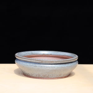 May include: A round, ceramic bonsai pot with a light blue exterior and a reddish-brown interior. The pot has a wide, shallow design with a slightly raised rim. The pot is sitting on a light-coloured surface against a black background.