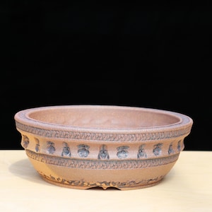 May include: A brown, round bonsai pot with a decorative band of repeating patterns. The pot has a wide, shallow design and is made of a textured clay material. The background is black.
