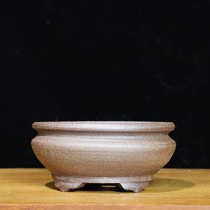 May include: A small, brown ceramic bonsai pot with a rounded shape and three small feet. The pot has a textured surface and a wide, shallow opening. The pot is designed for growing small plants and is suitable for indoor or outdoor use.