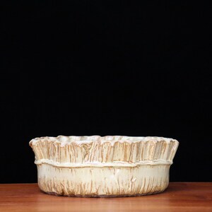 May include: A cream-colored, ceramic bowl with a textured, ruffled rim. The bowl sits on a wooden surface against a black background. The bowl's design features vertical lines and a band around the middle.