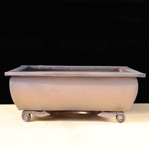 Puede incluir: Una maceta rectangular marrón para bonsái con lados redondeados y una parte superior plana. La maceta tiene pies decorativos con volutas y una superficie texturizada. El fondo es negro y la maceta está sobre una superficie de color claro.