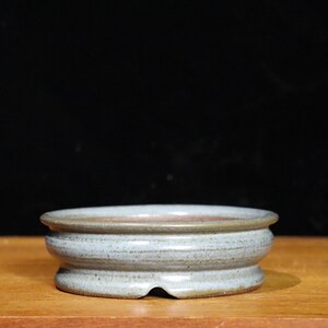 May include: A small, round, ceramic bonsai pot with a light blue-grey glaze. The pot has a wide, shallow design with a slightly raised rim and a drainage hole. The pot is sitting on a wooden surface.
