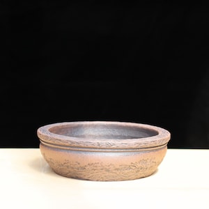 May include: A round, brown ceramic bonsai pot with a textured rim and a wave pattern around the sides. The pot has a slightly iridescent finish and sits on a white surface against a black background.