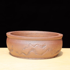 May include: A round, reddish-brown bonsai pot with a wide, shallow design. The pot features a textured surface with a carved, wave-like pattern around the exterior. The pot is made of ceramic and is suitable for bonsai trees.