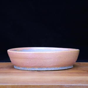 May include: A handmade, low-profile ceramic bowl with a light pink exterior and a grey base. The interior has a subtle blue and yellow hue. The bowl sits on a wooden surface against a black background. This bowl is ideal for serving food.