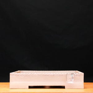 May include: A rectangular, pale-coloured box with a decorative, textured top edge. The box sits on a wooden surface against a dark background. The box has a small, rectangular cutout on one side.