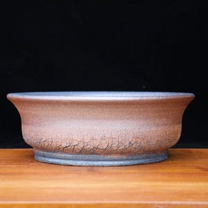 May include: A brown ceramic bonsai pot with a wide, shallow design. The pot has a textured surface with a cracked appearance and a dark rim. It sits on a wooden surface against a black background.