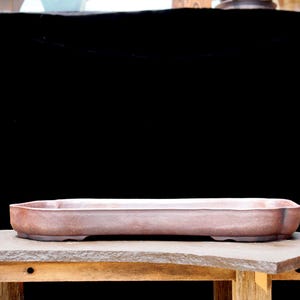 May include: A rectangular, reddish-brown bonsai pot with rounded corners and a low profile. The pot sits on a wooden table with a natural stone top, against a black background. The pot is designed for bonsai trees.