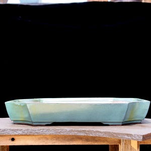 May include: A rectangular, pale blue ceramic bonsai pot with a geometric design. The pot has a wide, shallow shape with angled sides and a slightly raised base. The pot is displayed on a wooden surface against a black background.