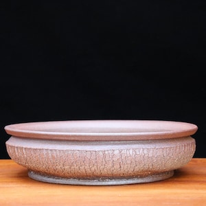 May include: A round, shallow bonsai pot with a textured, cracked surface. The pot is a muted reddish-brown color and has a wide, flat rim. The base is a lighter shade of the same color. The pot sits on a wooden surface.